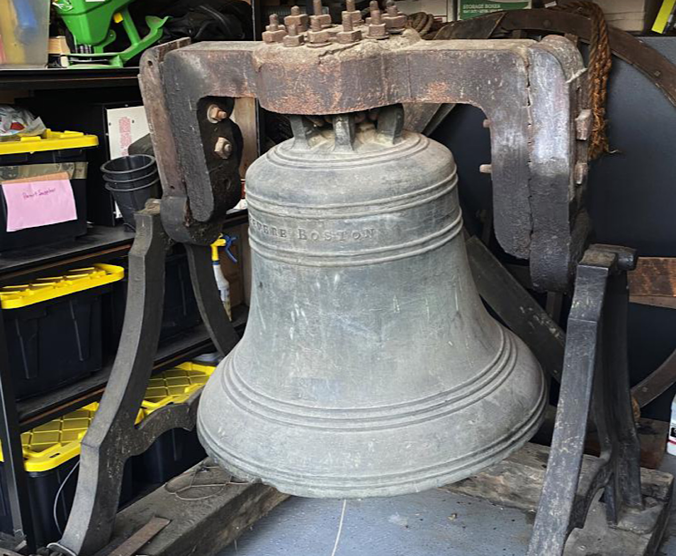 188-year-old bronze church bell rests on supports on the floor of a garage with clutter in the background