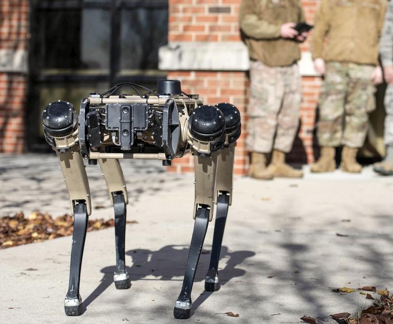 A robot dog with U.S. military airmen with a controller in the background.