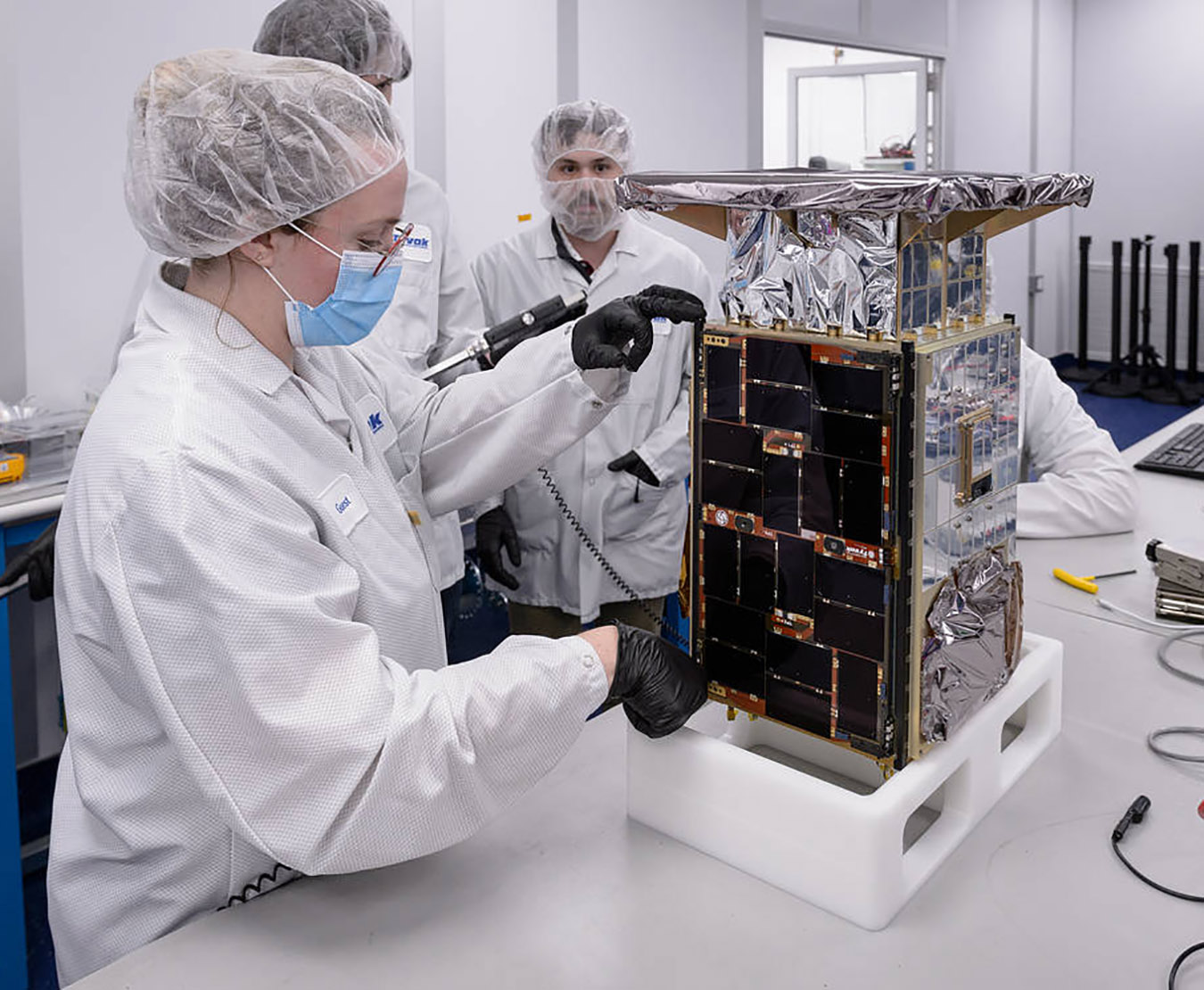 An engineer in a white lab suit holding a tape ruler to a small, microwave-sized, rectangular CAPSTONE satellite. One side has black panels, and others have silver panels and foil. Three team members are seen in the back.